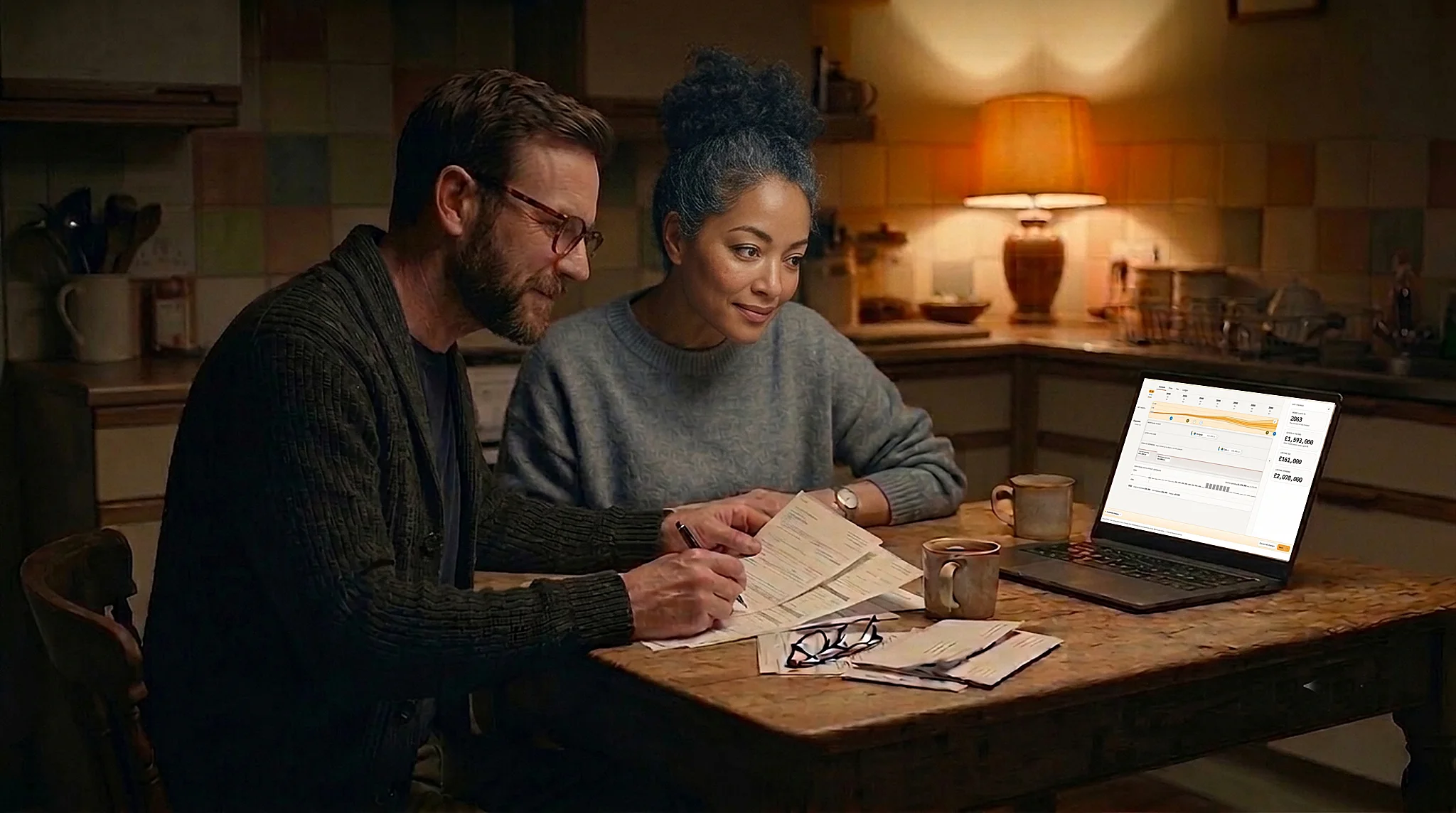 A couple at a kitchen table in the evening, reading paper pension and ISA statements beside a laptop showing a FutureClear projection.